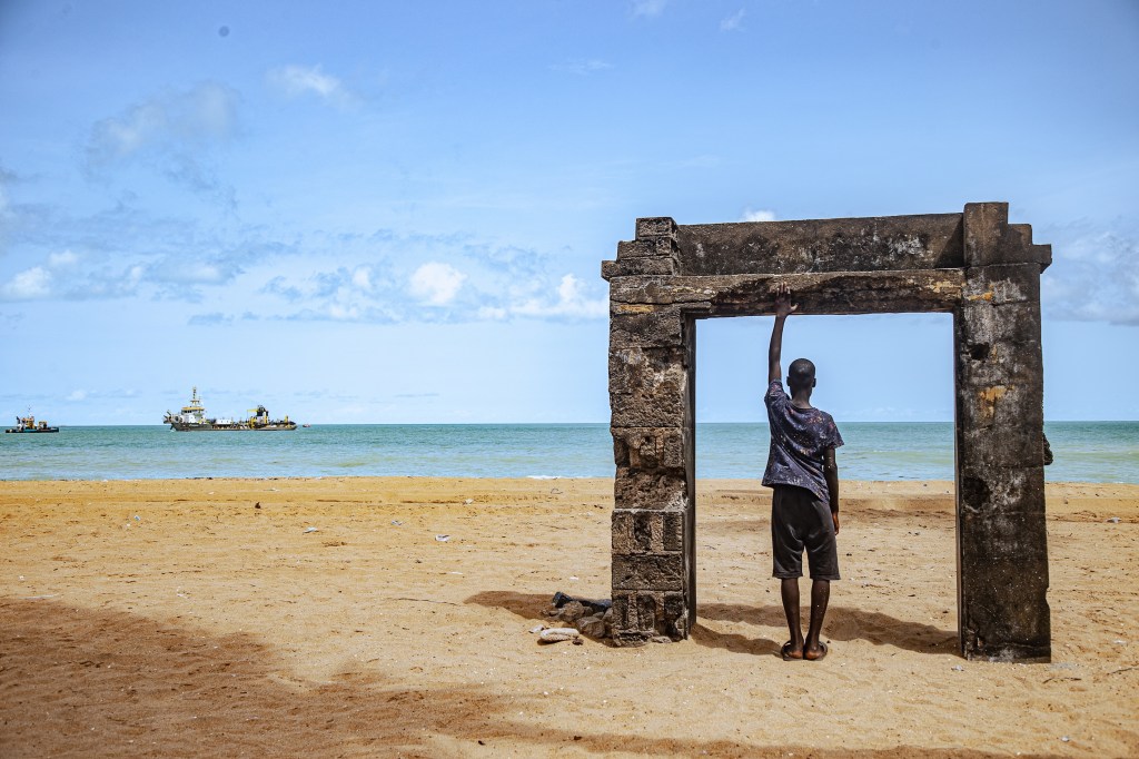 A child stands beneath a freestanding concrete doorway on a beach in Togo.