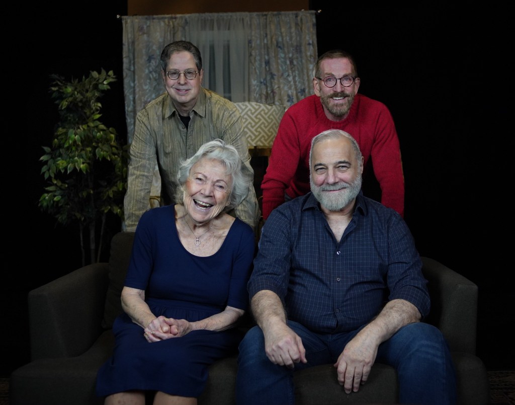 Four performers pose together in a theater setting, with two seated on a sofa and two standing behind them.