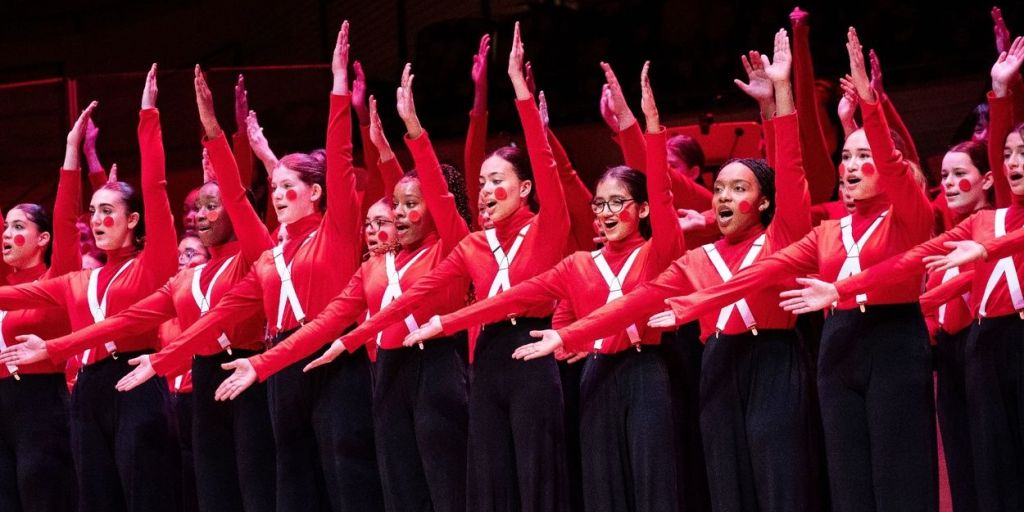 Young People's Chorus of New York City singers in red costumes performing onstage at a previous holiday concert.